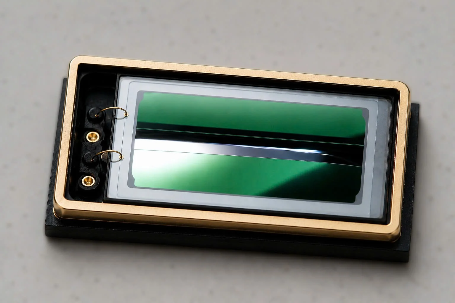 Macro photograph of the xMEMS Muir: a rectangular silicon MEMS die with a green-reflecting membrane, seated in a gold frame, with two gold wire-bond connections at the left edge.