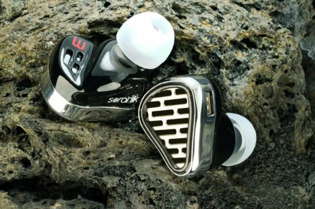 MEMS-Voix 3 in-ear monitors on dark volcanic rock: glossy black shells with polished metal faceplates, one showing a slotted grille, and white silicone eartips.