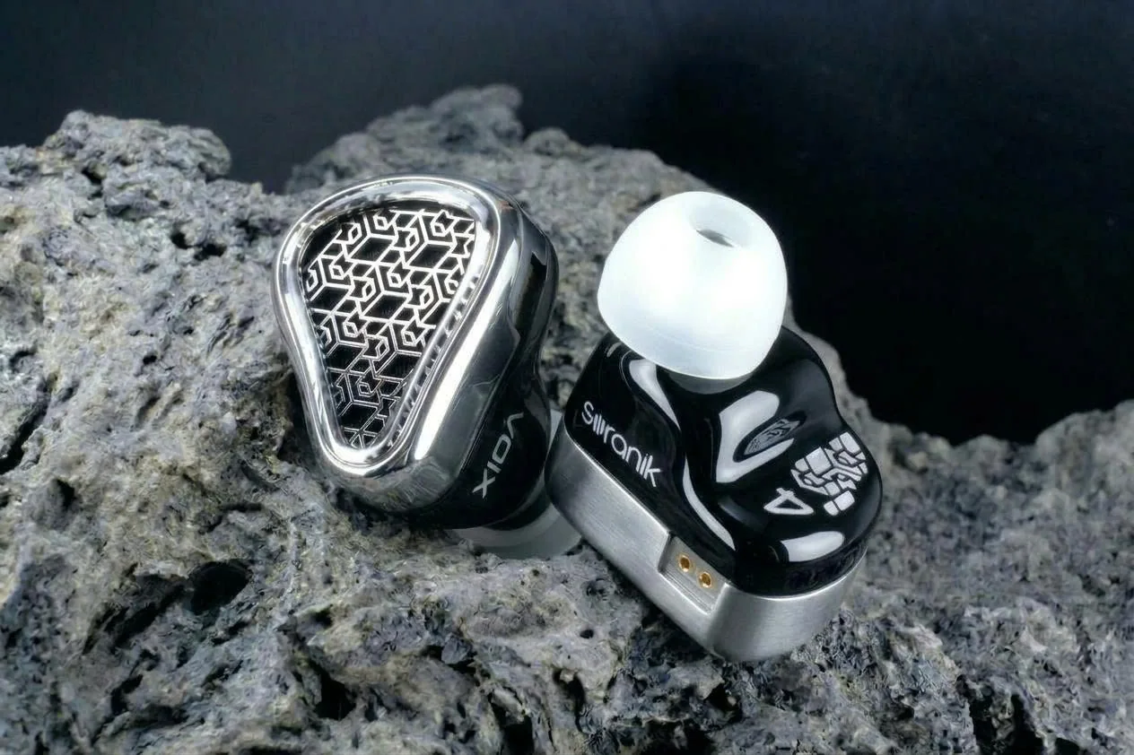 MEMS-Voix 4 in-ear monitors on dark volcanic rock: glossy black shell with a polished metal base and gold connector pins, and a mirror-finish faceplate with an engraved geometric pattern.